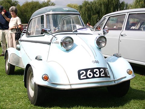 Head-on view of a pale blue bubble car with twin headlamps and a black plate, a white saloon and visitors beside it. Opens a larger view