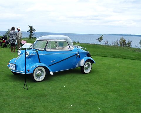 Blue bubble car with whitewall tyres on a lawn above a wide bay, a display sign on a stand by its nose. Opens a larger view