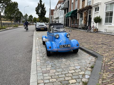 Bright blue bubble car with a dark hood parked on cobbles beside a brick pavement, old town houses behind. Opens a larger view