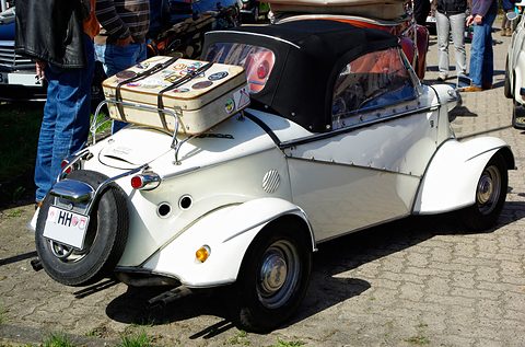 White soft-top bubble car from the rear left, a cream suitcase strapped to the luggage rack and a spare wheel below. Opens a larger view