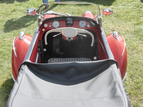 Looking into a narrow red cockpit: a cream handlebar-shaped steering yoke, two round dials and a checked seat, tonneau folded in front.
