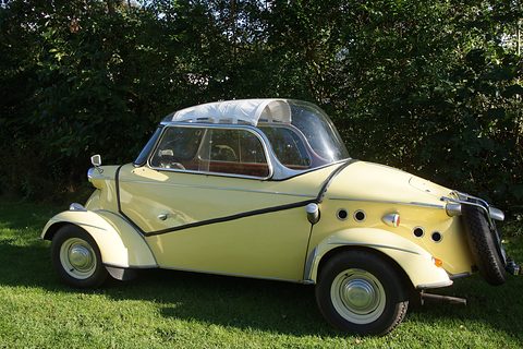 Side view of a cream bubble car with a clear canopy and dark side stripe, parked on grass in front of a hedge.