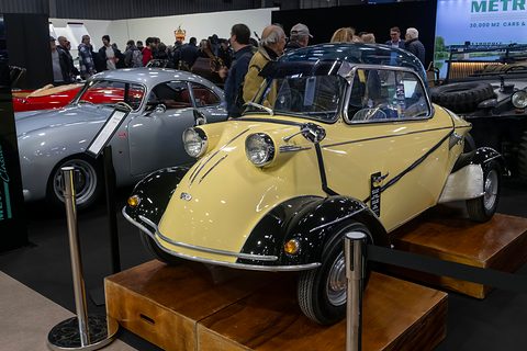 Cream bubble car with black front wings and a clear canopy on a wooden display plinth, visitors behind. Opens a larger view