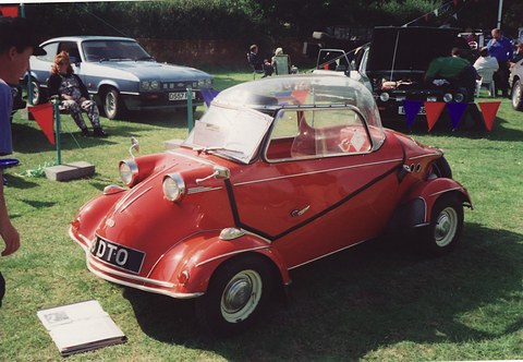 Red bubble car with a clear canopy and twin headlamps on its front wings, a show programme on the grass beside it. Opens a larger view