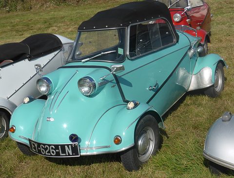 Turquoise bubble car with a black fabric top seen from the front left on grass, other microcars parked around it. Opens a larger view