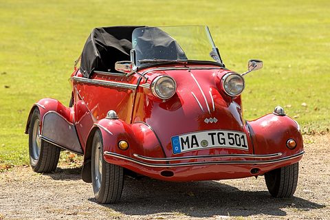 Red open-topped bubble car with a folded black hood and windscreen, twin headlamps on its wings, on a gravel path by a lawn. Opens a larger view