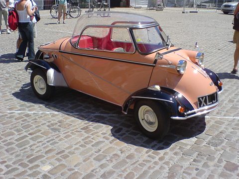 A four-wheeled tandem bubble car in salmon and black with a clear Plexiglas canopy, people walking behind. Opens a larger view