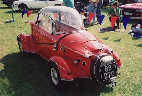 Red tandem-seat bubble car on grass seen from behind, spare wheel with plate 88 DTO on the tail, bunting and visitors behind. Opens a larger view