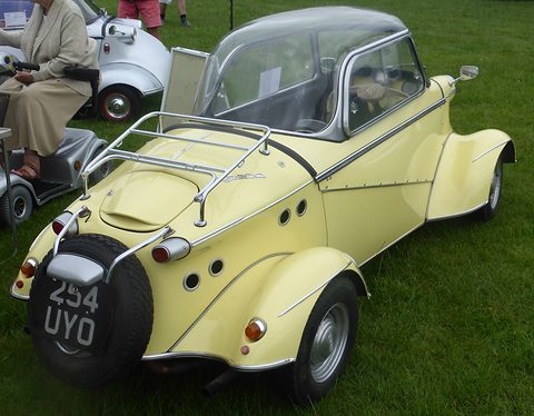 Rear quarter of a pale yellow tandem-seat car with a chrome luggage rack, spare wheel on the tail and round cooling holes. Opens a larger view