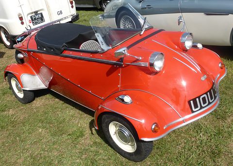 Red open two-seater with a low windscreen, black tonneau and twin headlamps on swelling front wings, on grass. Opens a larger view