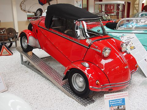 Red tandem two-seater with a black fabric hood on a chequer-plate ramp, a turquoise car and display boards behind. Opens a larger view