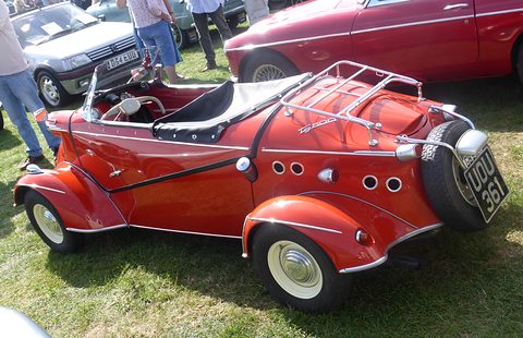 Side view of a red open tandem two-seater with a black tonneau, chrome luggage rack and spare wheel on the tail. Opens a larger view