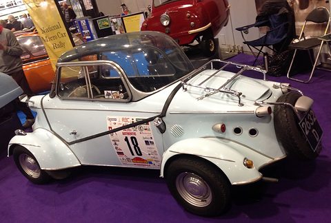 White bubble car with a canopy and rally number 18 on its door, luggage rack on the tail, on a purple show carpet. Opens a larger view