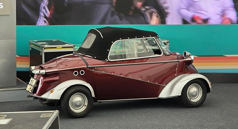 Side view of a maroon bubble car with a cream lower body, black fabric hood and whitewall tyres, in front of a display backdrop. Opens a larger view