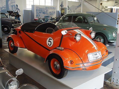 Orange open two-seater with a white stripe and a black number 5 roundel on a white museum plinth. Opens a larger view