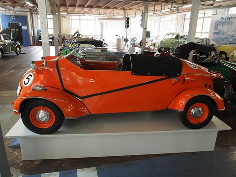 Side view of an orange open tandem two-seater with a black tonneau and a low screen on a white plinth in a factory hall. Opens a larger view