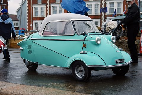 Turquoise bubble car with a white canopy roof parked on a wet road, red-brick houses and visitors with umbrellas behind. Opens a larger view
