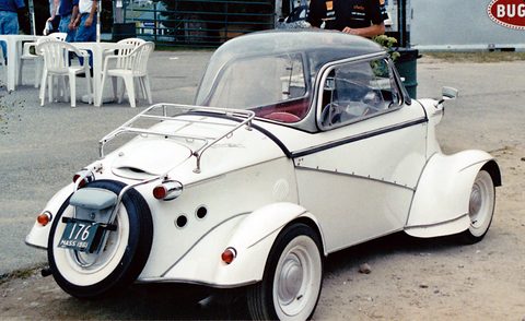 Rear quarter of a white bubble car with a clear canopy, luggage rack and spare wheel on the tail, café chairs behind. Opens a larger view