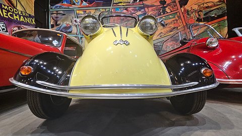 Wide-angle front view of a yellow bubble car with black wings, the FMR diamond badge and a chrome bumper, comic-strip murals behind. Opens a larger view