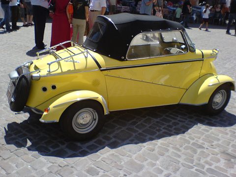 Rear three-quarter view of a yellow four-wheeled tandem car with a black fabric hood, chrome rack and tail-mounted spare. Opens a larger view
