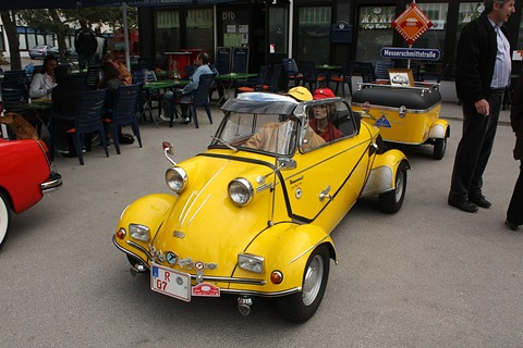 Yellow open tandem two-seater with two helmeted occupants, a matching yellow single-axle trailer behind, café tables in the background. Opens a larger view