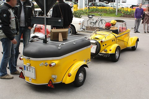 Yellow two-wheeled trailer with a black lid seen from behind, hitched to a yellow open Tg500, onlookers at the left. Opens a larger view