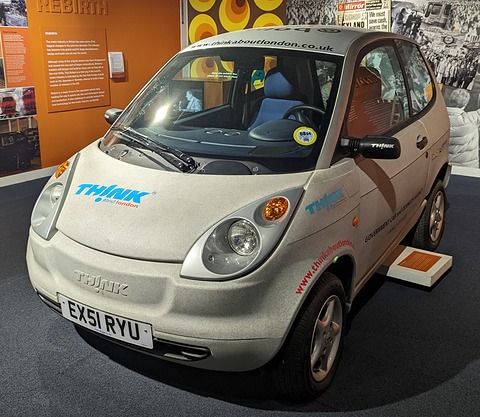 Cream small two-door car with blue TH!NK decals, round headlamps and an EX51 plate on a dark museum floor. Opens a larger view