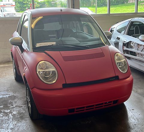 Matt red small hatchback with round headlamps and a dark grille slot on a wet concrete floor by large windows.