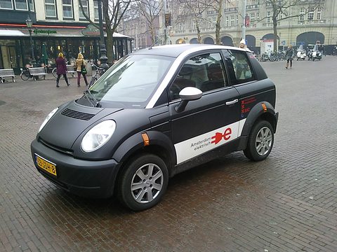 Black small hatchback with a white door panel and red plug logo on brick paving, a hotel facade behind. Opens a larger view