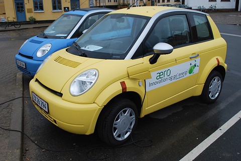 Yellow small hatchback with ‘aero’ door lettering beside a blue one, both plugged in on a wet car park. Opens a larger view