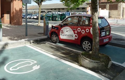 Red and white small hatchback with Carsharing lettering parked by a charging post, an electric-car symbol painted on the bay. Opens a larger view