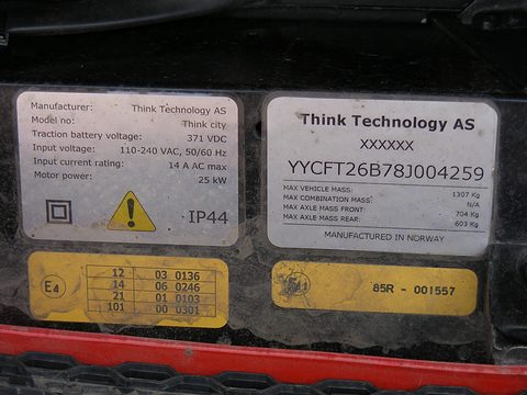Two white printed maker’s plates and two yellow approval labels screwed to a black panel above a red bumper. Opens a larger view