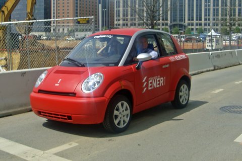 Red small hatchback with white ENER1 lettering and a driver at the wheel on a road beside building works. Opens a larger view
