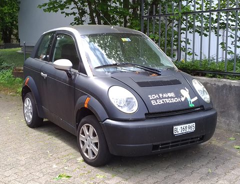 Matt black small hatchback with round headlamps and a Swiss number plate on paving beside an iron fence. Opens a larger view