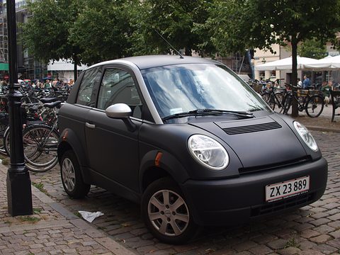 Matt black small hatchback with round headlamps and a Danish plate on cobbles beside rows of parked bicycles. Opens a larger view