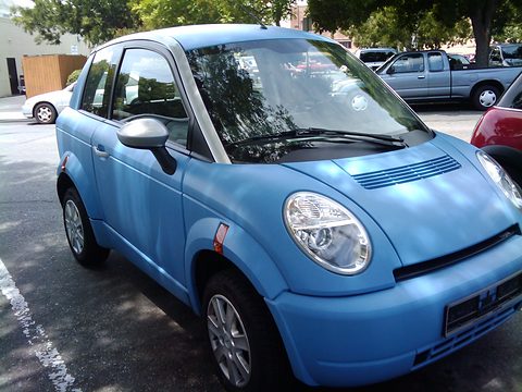 Light-blue small hatchback with large round headlamps and bonnet vents, parked in shade beside pick-up trucks.