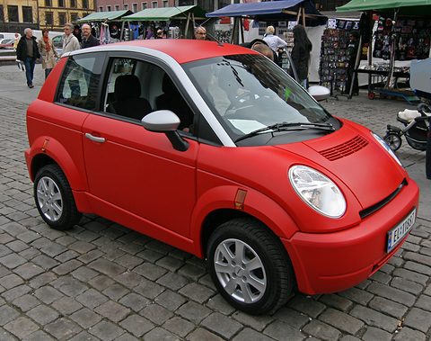 Red small hatchback with round headlamps and bonnet vents on cobblestones, market stalls and shoppers behind. Opens a larger view