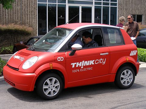 Red small hatchback lettered THINK city with a man at the wheel and visitors in front of an office. Opens a larger view