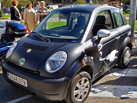 Black small hatchback with a white plug-and-cable graphic on its door and a Spanish plate, parked by a kerb. Opens a larger view