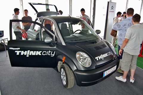 Black small hatchback lettered THINK city with both doors open, visitors looking at it on a stand. Opens a larger view