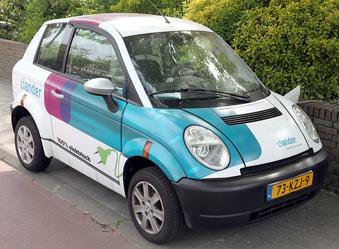 White small hatchback with teal and purple panels and a yellow Dutch plate parked on a pavement by a hedge. Opens a larger view