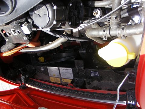 Engine bay crowded with an electric motor housing, orange high-voltage cables, a coolant bottle and labels. Opens a larger view