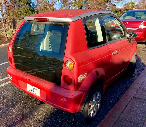 Rear of a red hatchback with a black glass tailgate, round tail lamps and a white plate in low winter sun. Opens a larger view