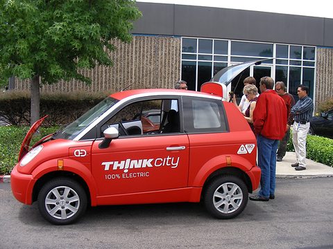 Side view of a red hatchback lettered THINK city, its tailgate raised, a group of visitors at the back. Opens a larger view