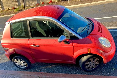 Red small hatchback from above and to the side, its silver roof frame and windscreen catching the sun at a kerb. Opens a larger view