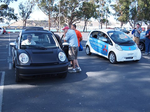 Black small hatchback with its driver inside next to a white egg-shaped electric car, people talking between them. Opens a larger view