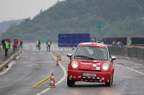 Red small hatchback with chequered rally livery driving between traffic cones on a wet road, marshals behind. Opens a larger view