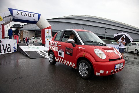 Red and white small hatchback with rally number 01 beside an inflatable START arch, people with umbrellas behind.