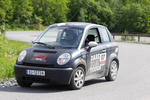 Black small hatchback with rally number 07 and a helmeted driver cornering on a grey track lined with trees. Opens a larger view