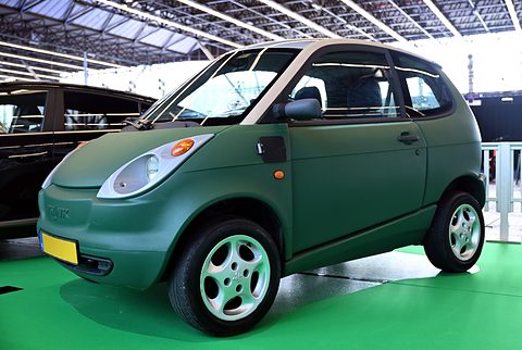 Matt green small two-door car with round headlamps and grey wheels on green carpet under a glass roof. Opens a larger view